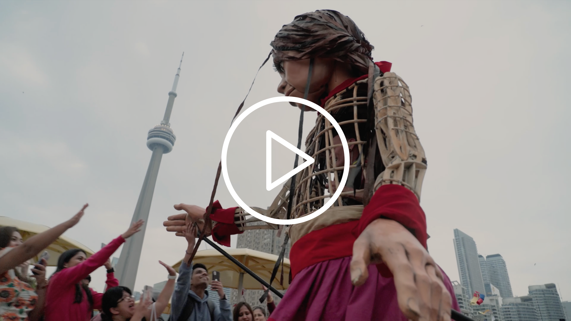 Hyperlinked thumbnail from our festival highlight reel, watchable on YouTube. The image is a view of Amal from below, greeting people in Toronto. The CN tower is in the background. A white play button (an outlined circle with an outlined triangle inside) sits on top of the image in the middle.