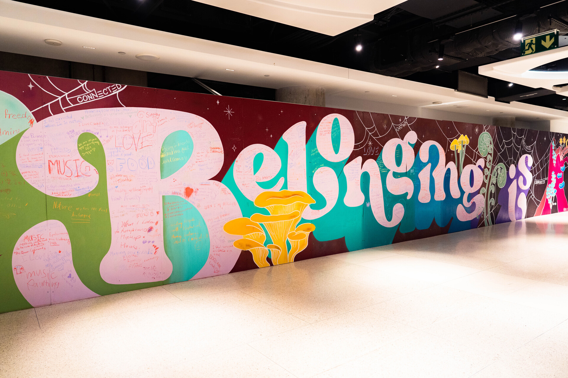 Photo of Belonging Is at the Union Station Foodie Aisle by Cassandra Popescu. The words Belonging Is is in big curly white font on a long wall. Embellished with colourful mushrooms and cobwebs. A lot of space is filled with written entries from passerbys.